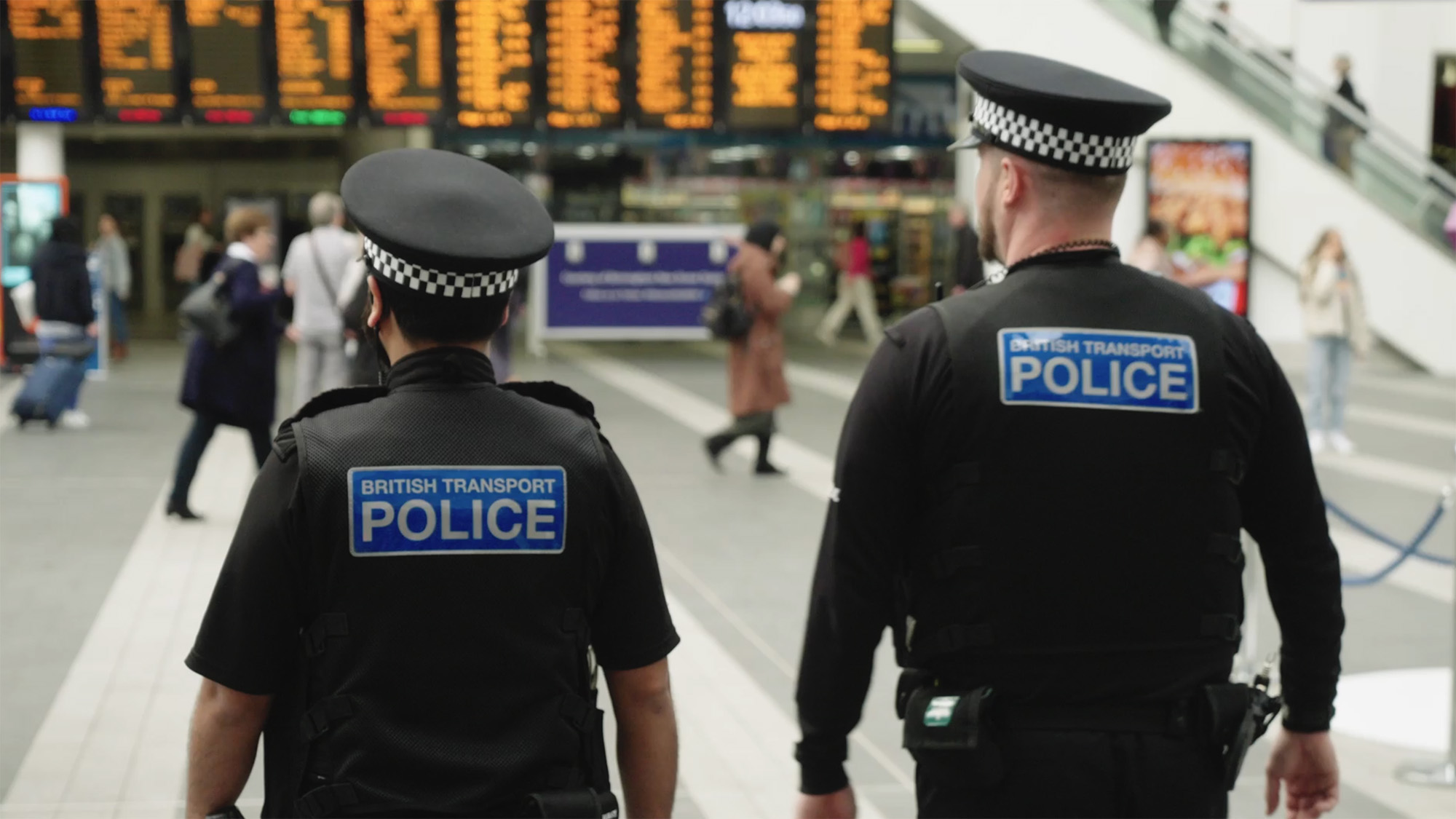 British Transport Police officer standing in a busy train station with departure boards and passengers in the background