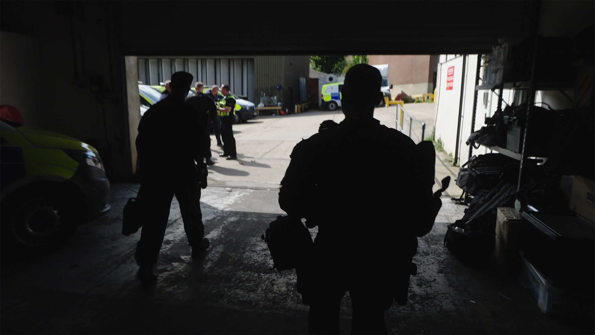 Uniformed officers walking from a dimly lit garage toward police vehicles during an operational briefing