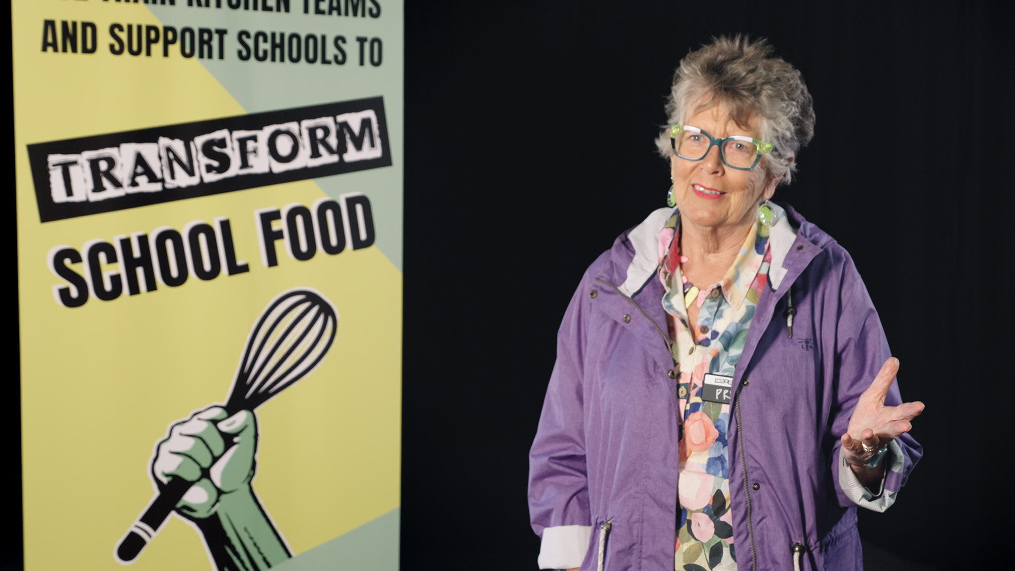 Dame Prue Leith standing beside a promotional banner for a school food programme, wearing colourful clothing and glasses