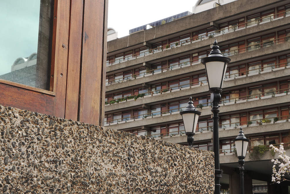 Brutalist concrete building with a wooden‑framed window in the foreground and multi‑storey balconies with greenery in the background