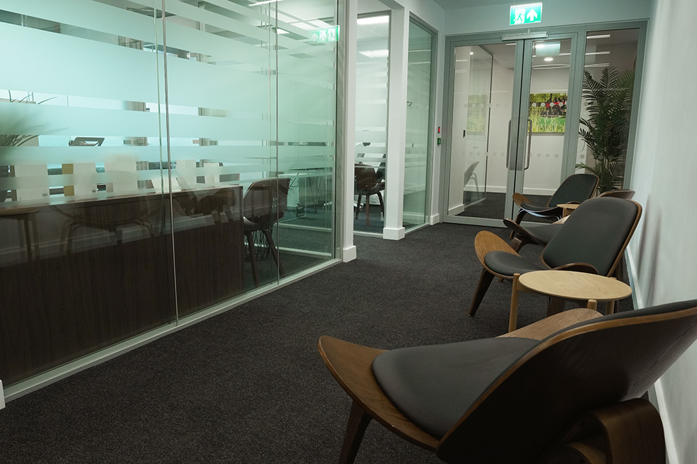 Modern office corridor with wooden chairs, frosted‑glass meeting room and double doors leading to a workspace