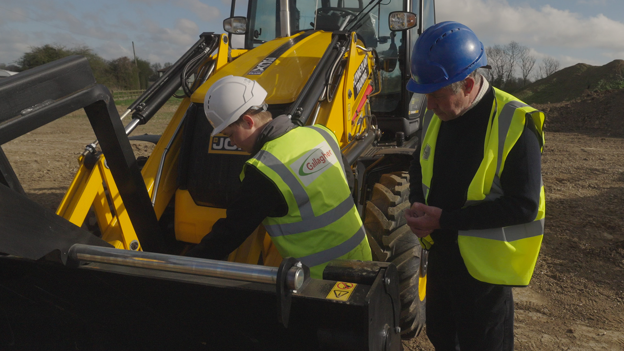 Finn Gallagher and Derek Mansfield in safety gear inspecting the front bucket of a yellow JCB backhoe loader on a construction site