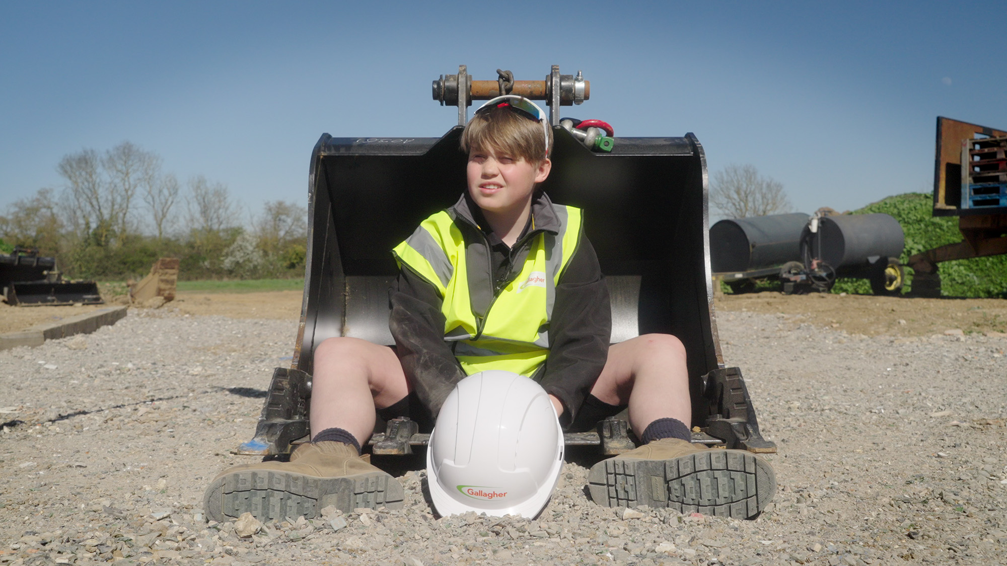 Finn Gallagher in high‑visibility vest sitting in an excavator bucket with a Gallagher hard hat placed on the ground