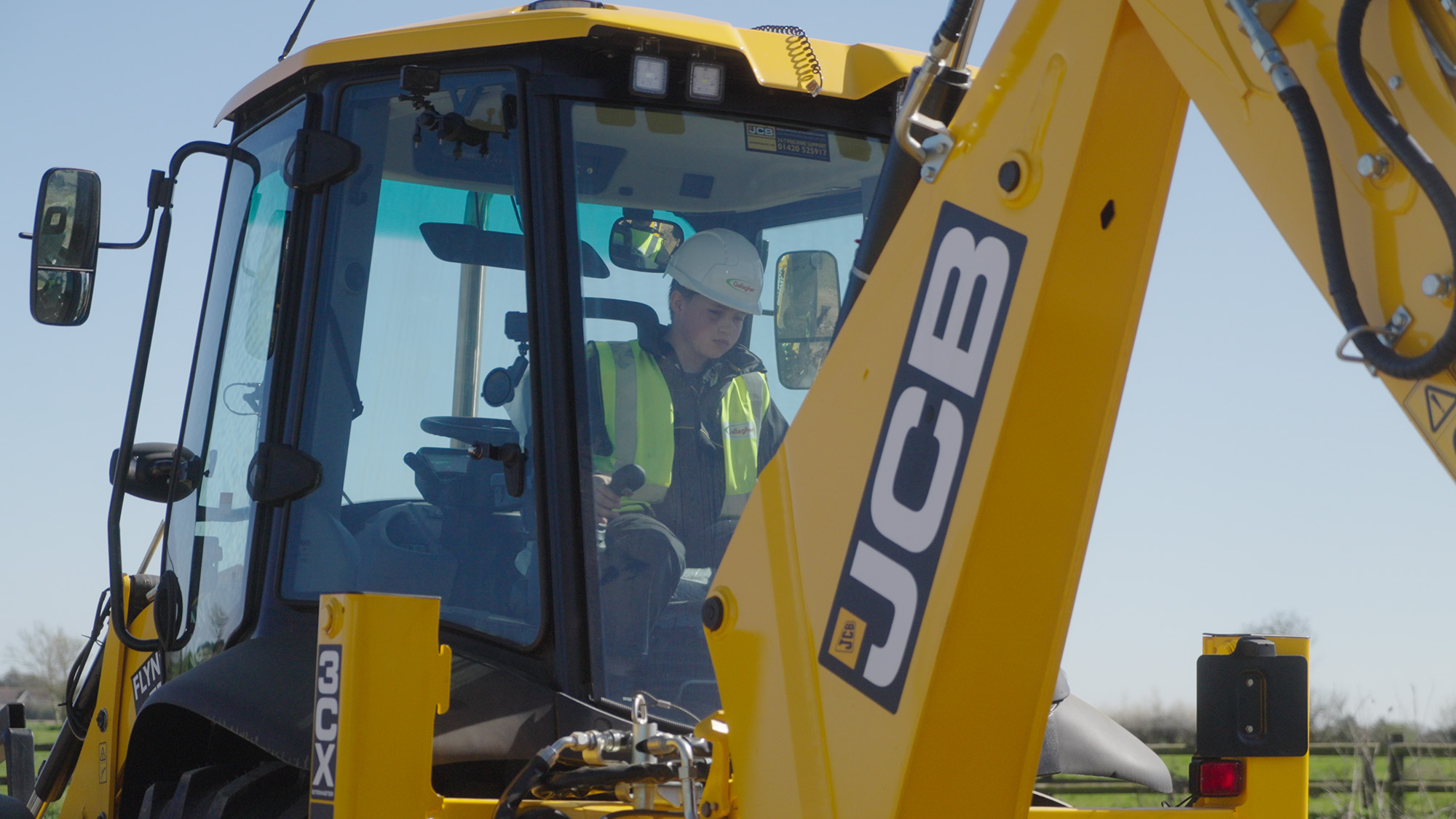 Finn Gallagher wearing a safety helmet and high‑visibility vest seated inside the cab of a JCB 3CX backhoe loader