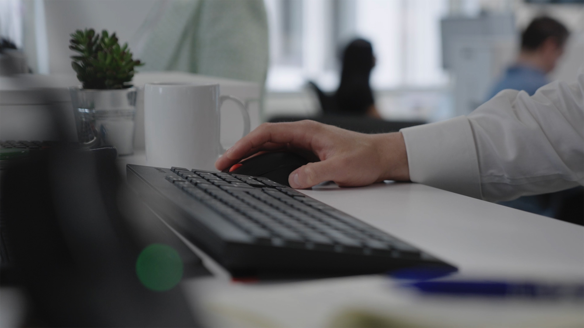 A close‑up shot of a hand using a computer mouse beside a keyboard on a white desk, with a coffee mug, plant and pen nearby. Blurred office workers sit in the background