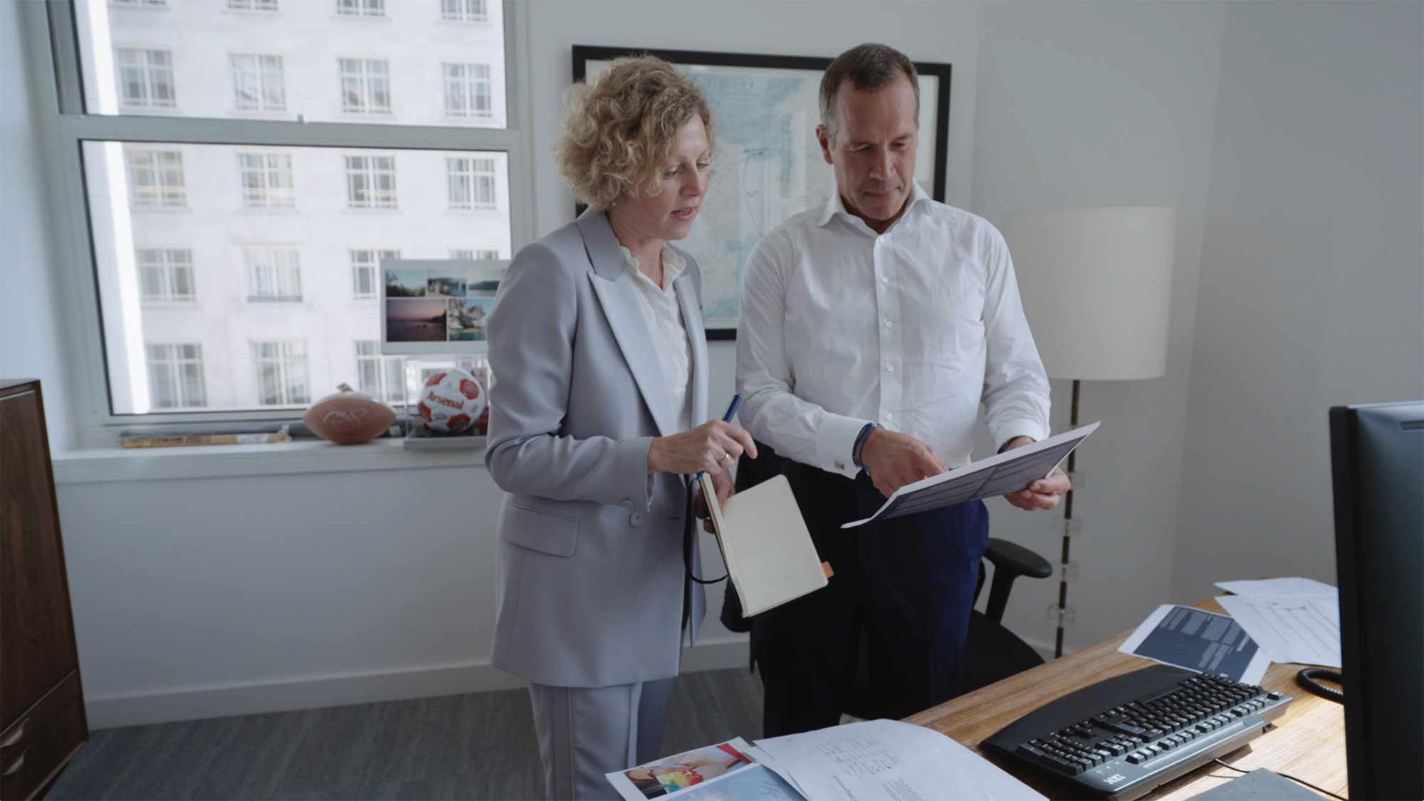Two people stand in an office reviewing papers — one holding a notebook and pen, the other holding a printed sheet. The workspace includes a desk with documents, a computer monitor, and personal items such as photos and a football