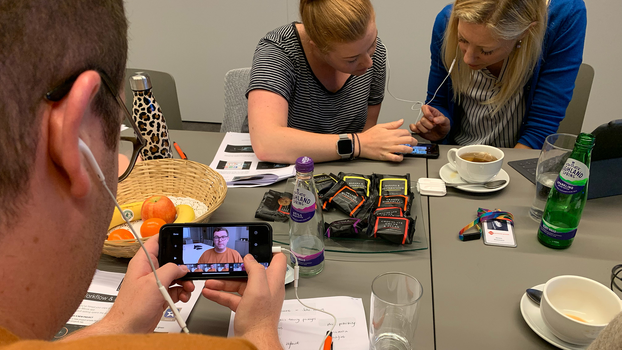 Group using smartphones and earphones around a table with fruit, drinks, notes, and a laptop during a collaborative session