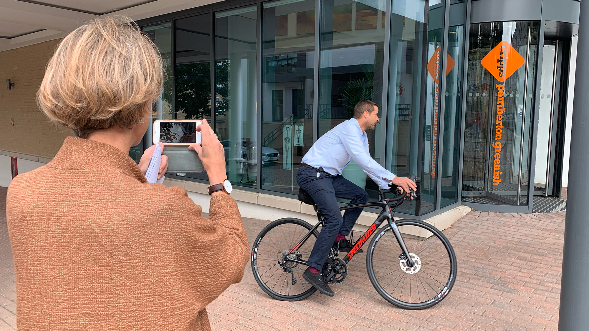 Person taking a video of a cyclist riding past a modern glass building with signage
