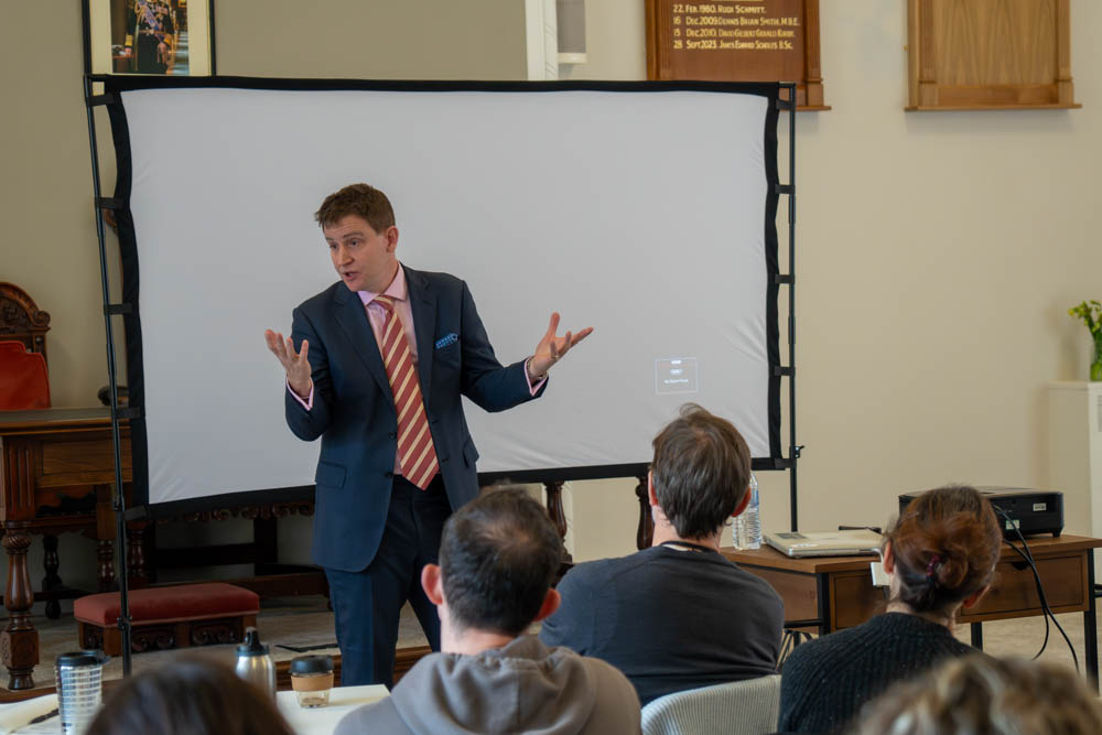 Mike Martin MP in a suit delivering a presentation to attendees seated at tables in a training room