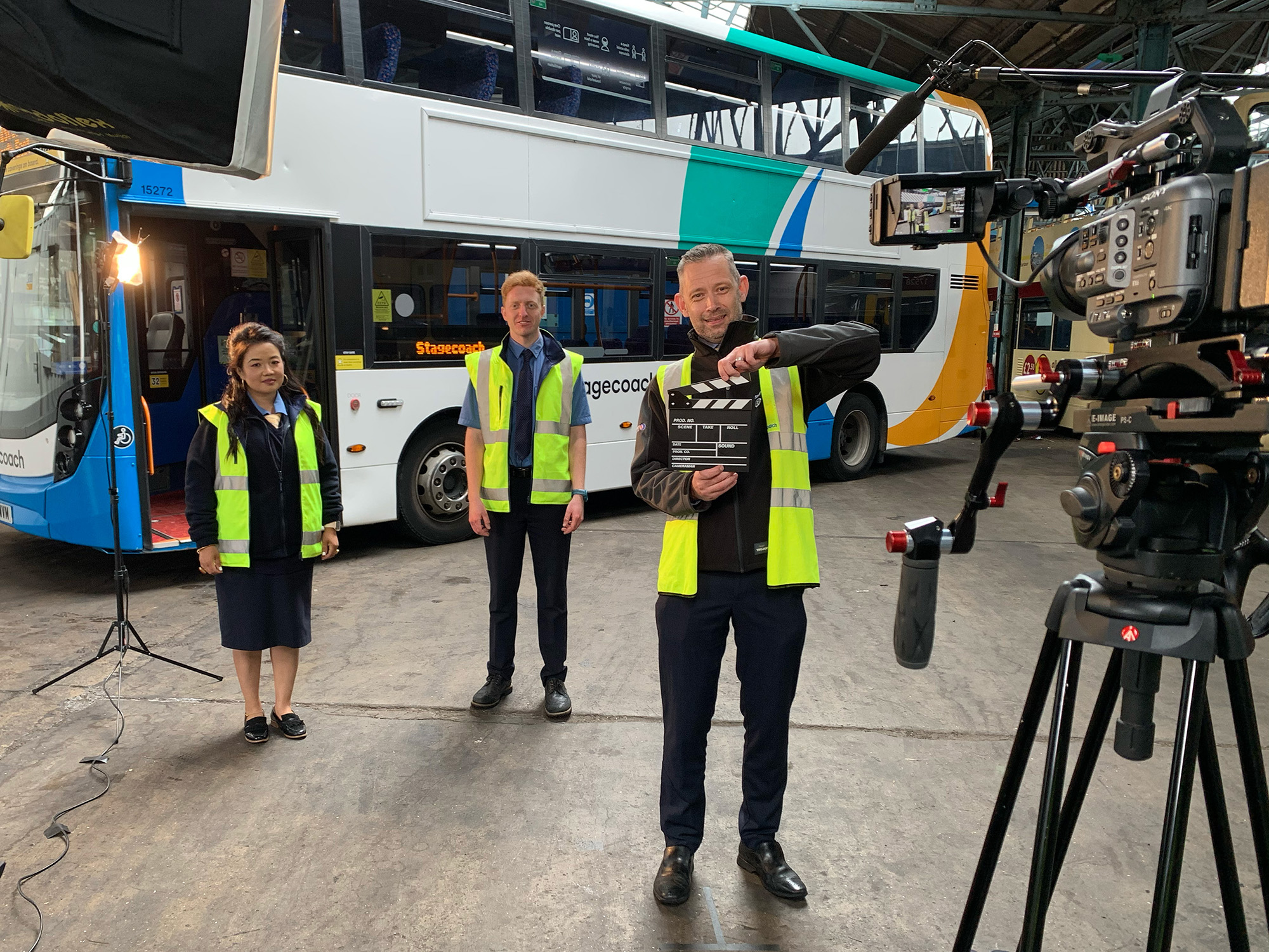 Film crew recording three people in high‑vis vests in front of a Stagecoach double‑decker bus