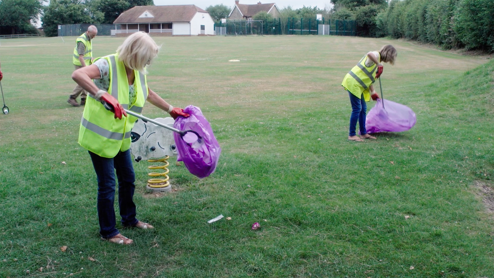 Volunteers in high‑visibility vests collecting litter in a grassy park area with a playground nearby