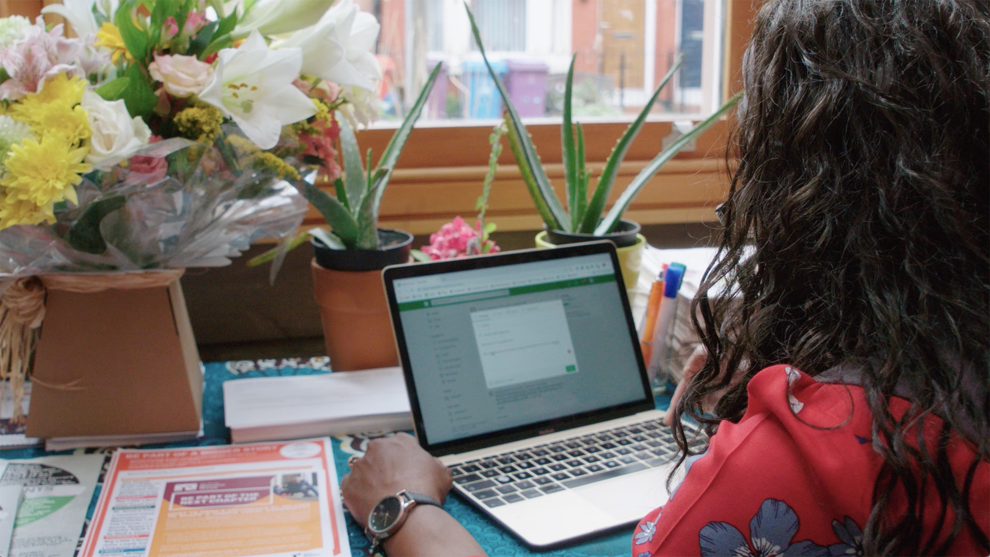 Person typing on a laptop at a table with papers and houseplants beside a window overlooking a residential street