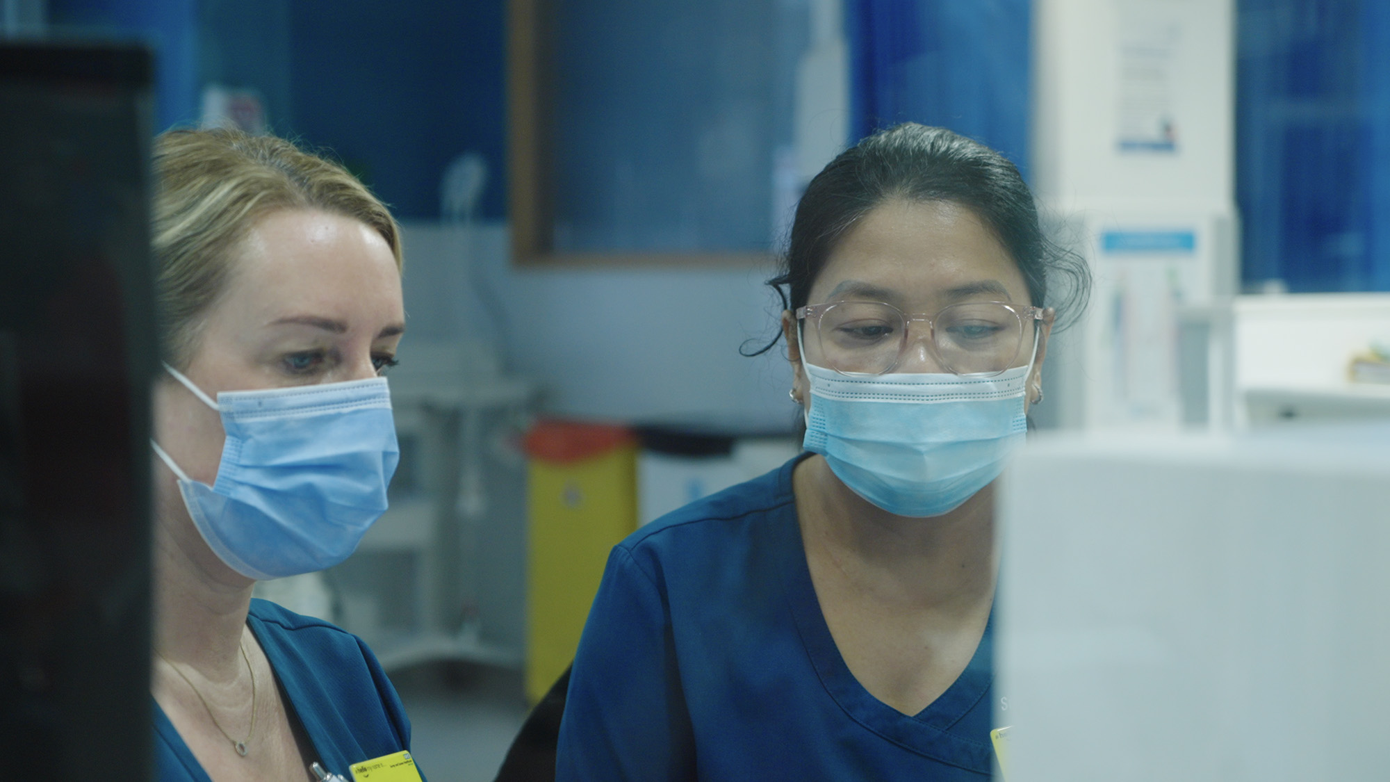 Two healthcare workers in blue scrubs and masks reviewing information on a computer in a clinical setting