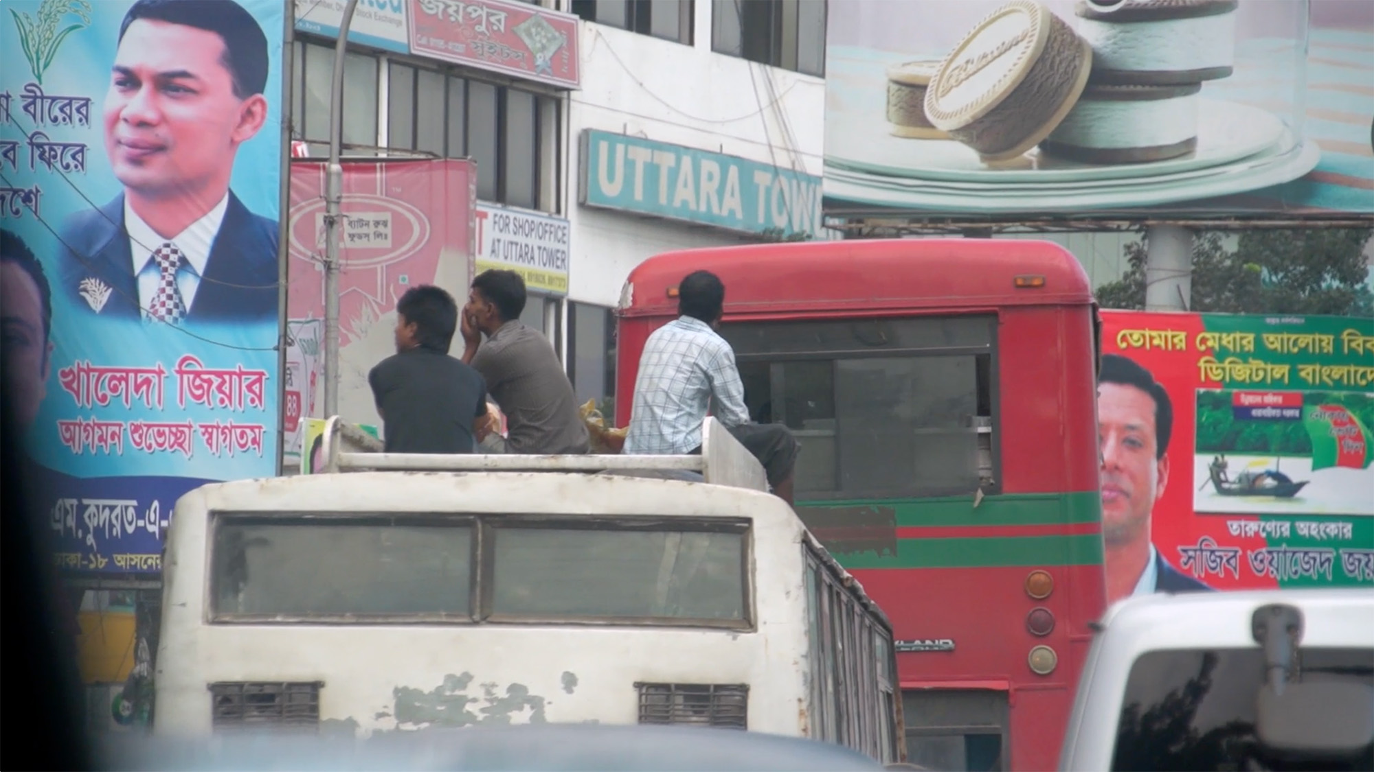 Crowded urban street in Dhaka with vehicles, people on a truck, and Bengali advertising billboards