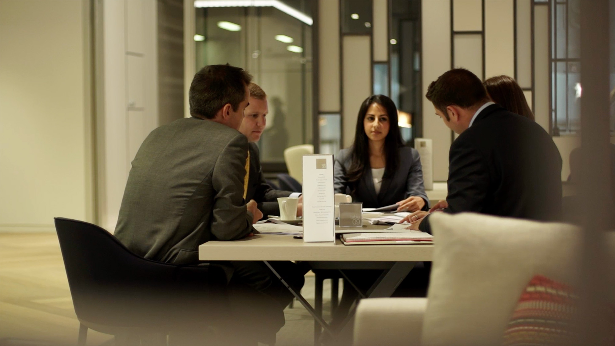 Five professionals seated around a table in a modern office discussing documents and notes