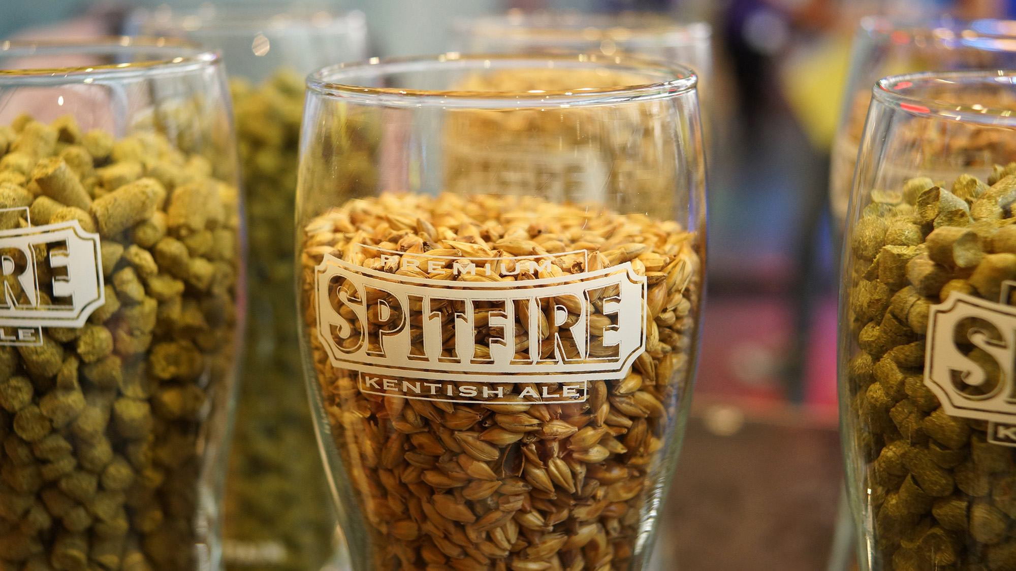 Glasses filled with malted barley and hop pellets, including one branded Spitfire Kentish Ale