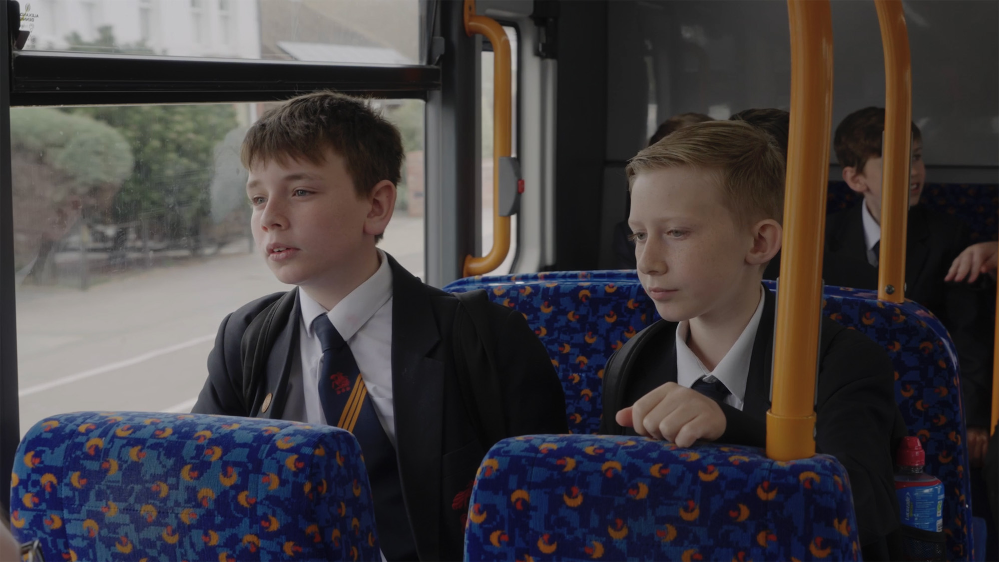 Two students in school uniform seated on a bus with patterned blue seats
