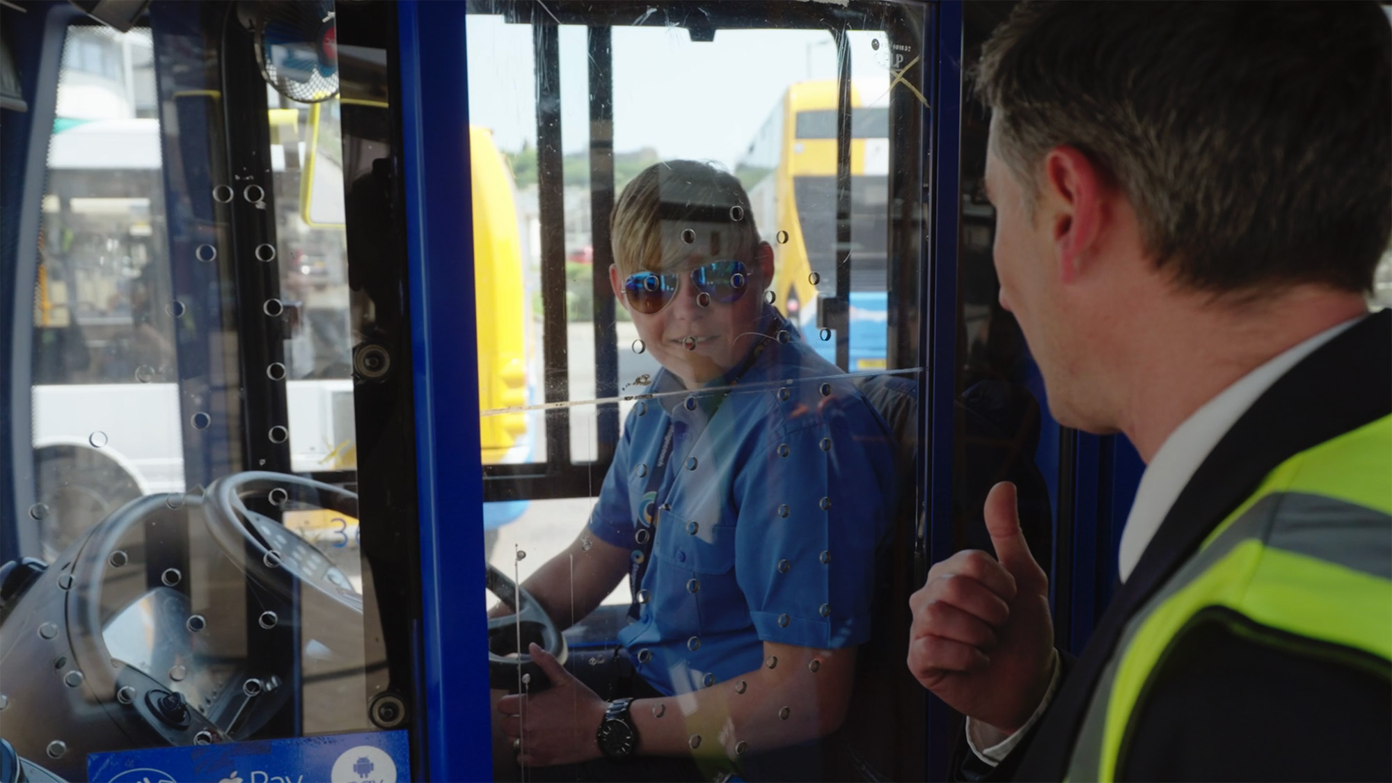 Bus driver behind a protective screen giving a thumbs‑up while another worker responds outside the cab