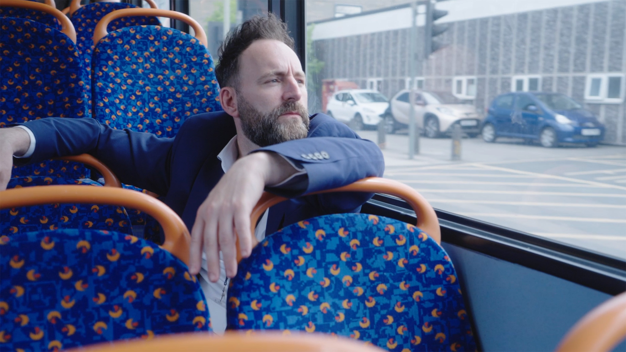 Joel Mitchell in a suit seated on a bus with patterned blue seats, looking out of the window