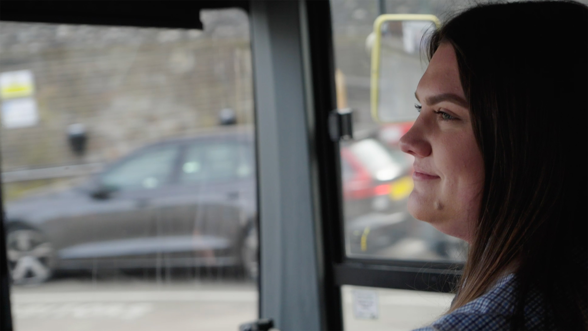 Bus driver with long brown hair looking out of a vehicle window at passing cars and a stone wall