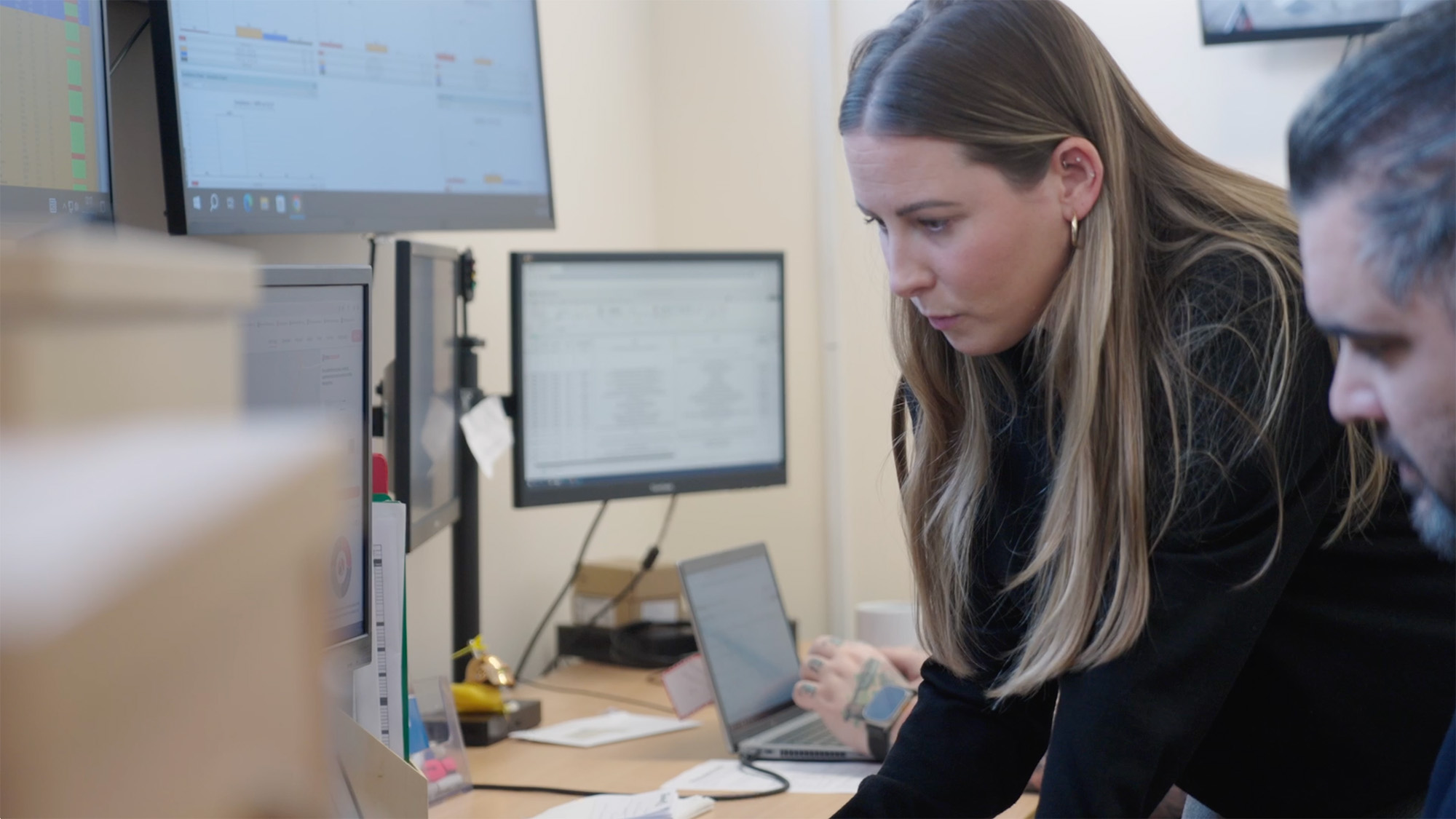 Two people working at desks with multiple monitors displaying spreadsheets and charts