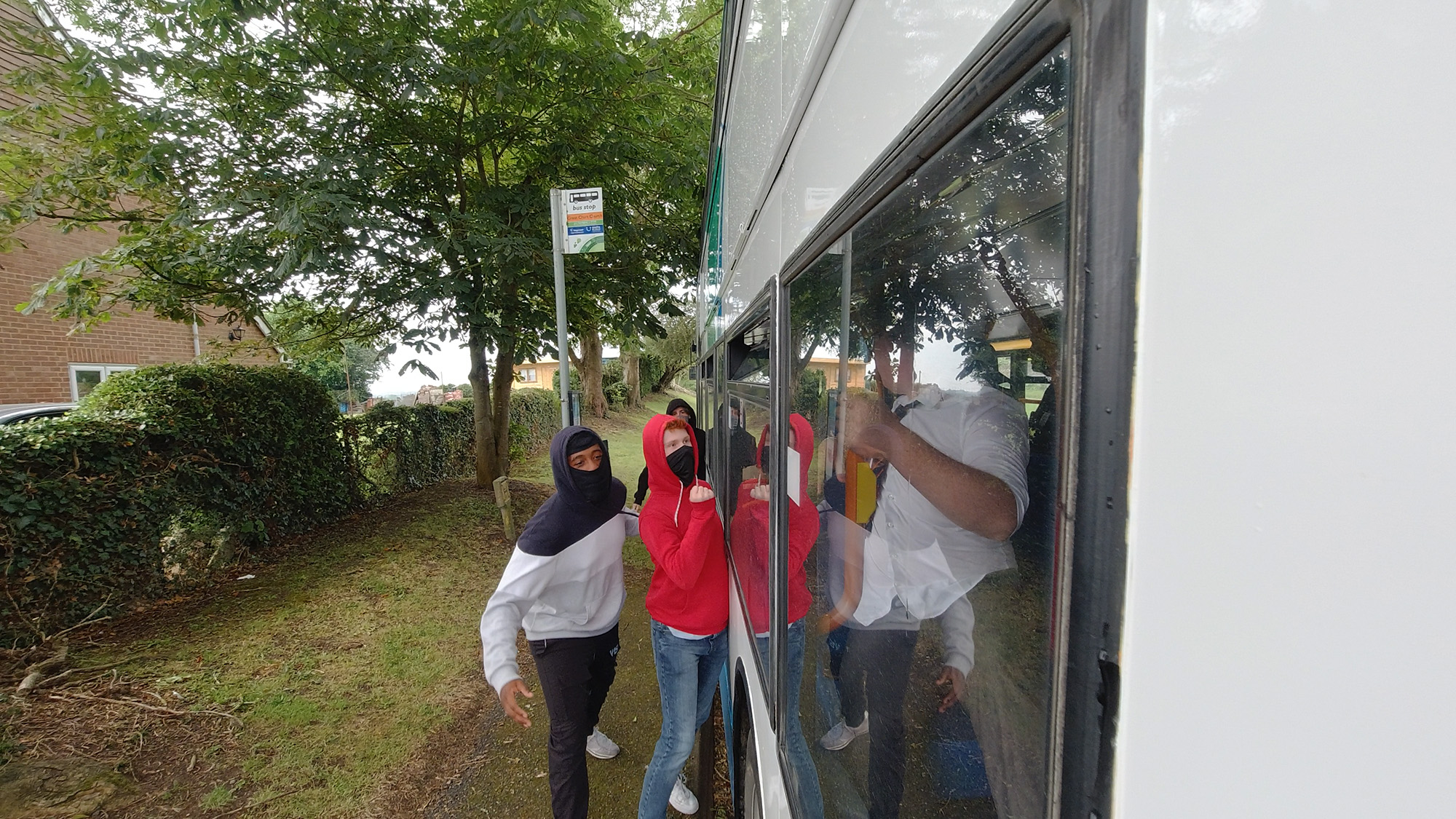 Three hooded people with face coverings at a bus stop next to a bus, with one person visible through the window