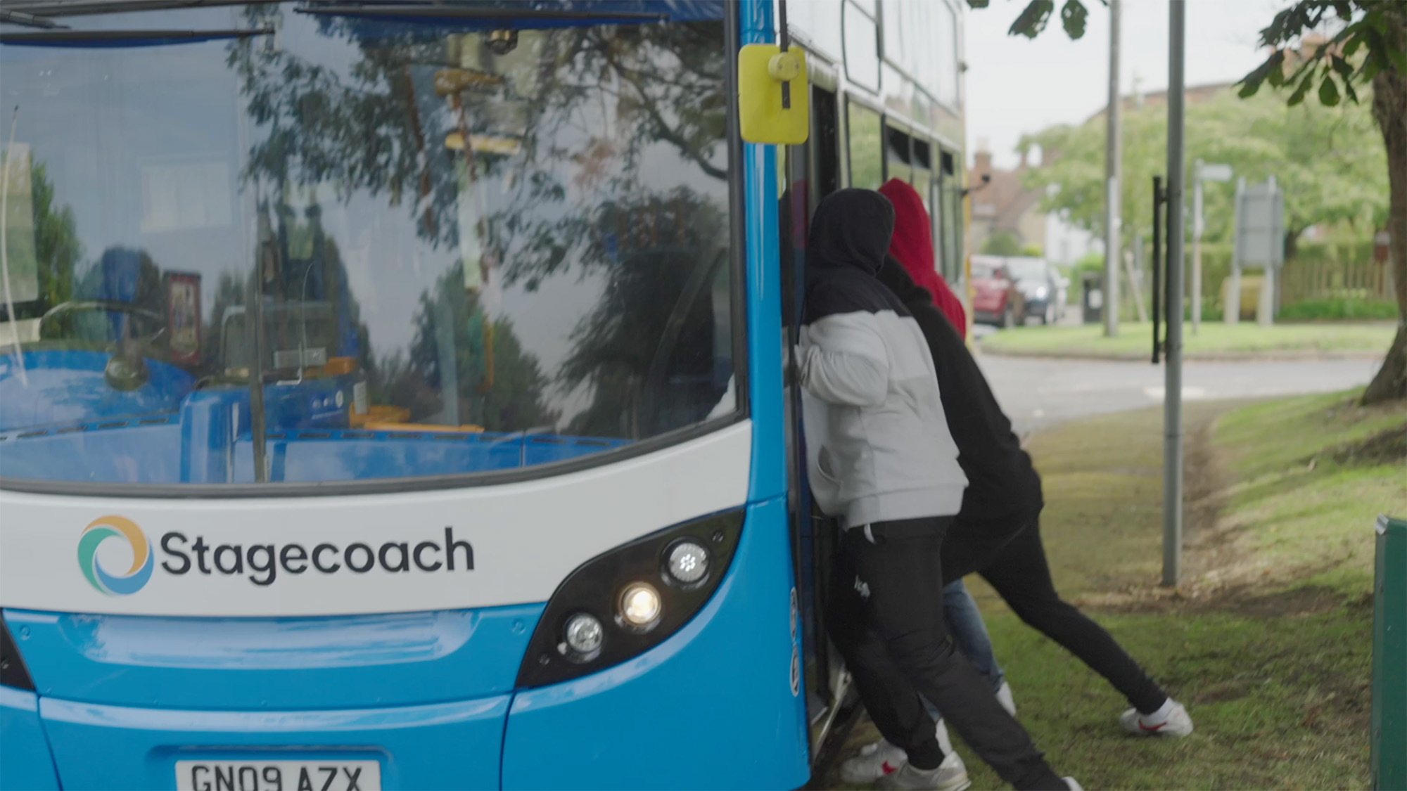 Three people in hoodies pushing against the front of a Stagecoach bus on a grassy roadside