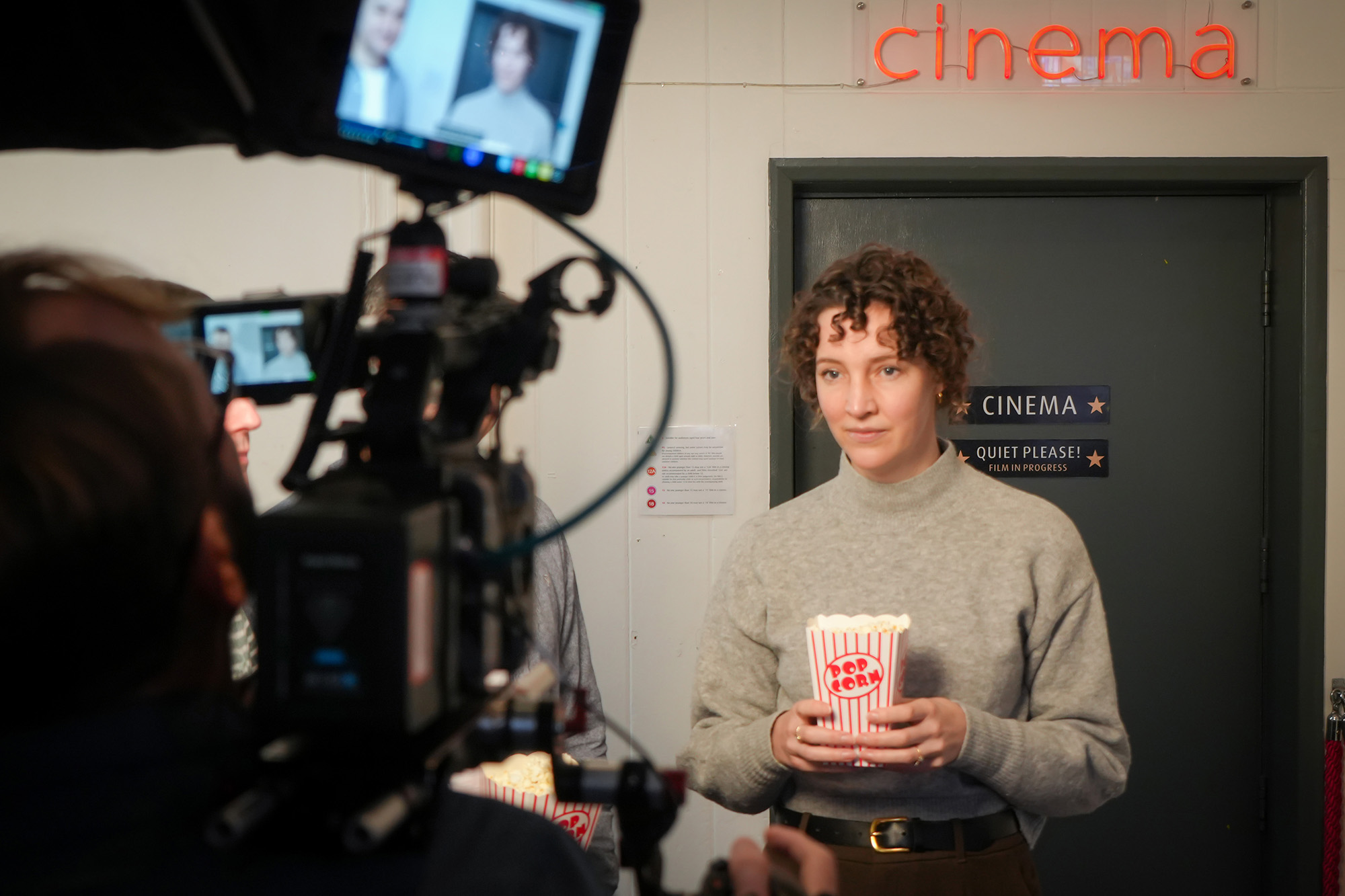 Camera crew filming a person holding popcorn outside a cinema door with signage