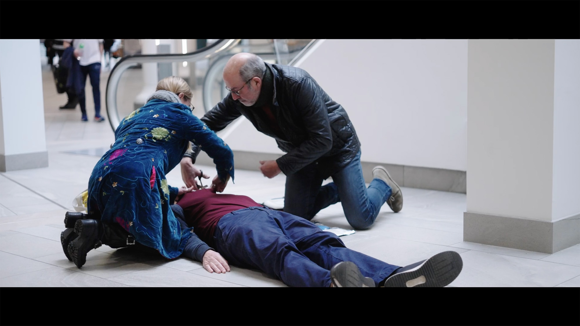 Two people kneeling to assist someone lying on the floor during a first‑aid emergency