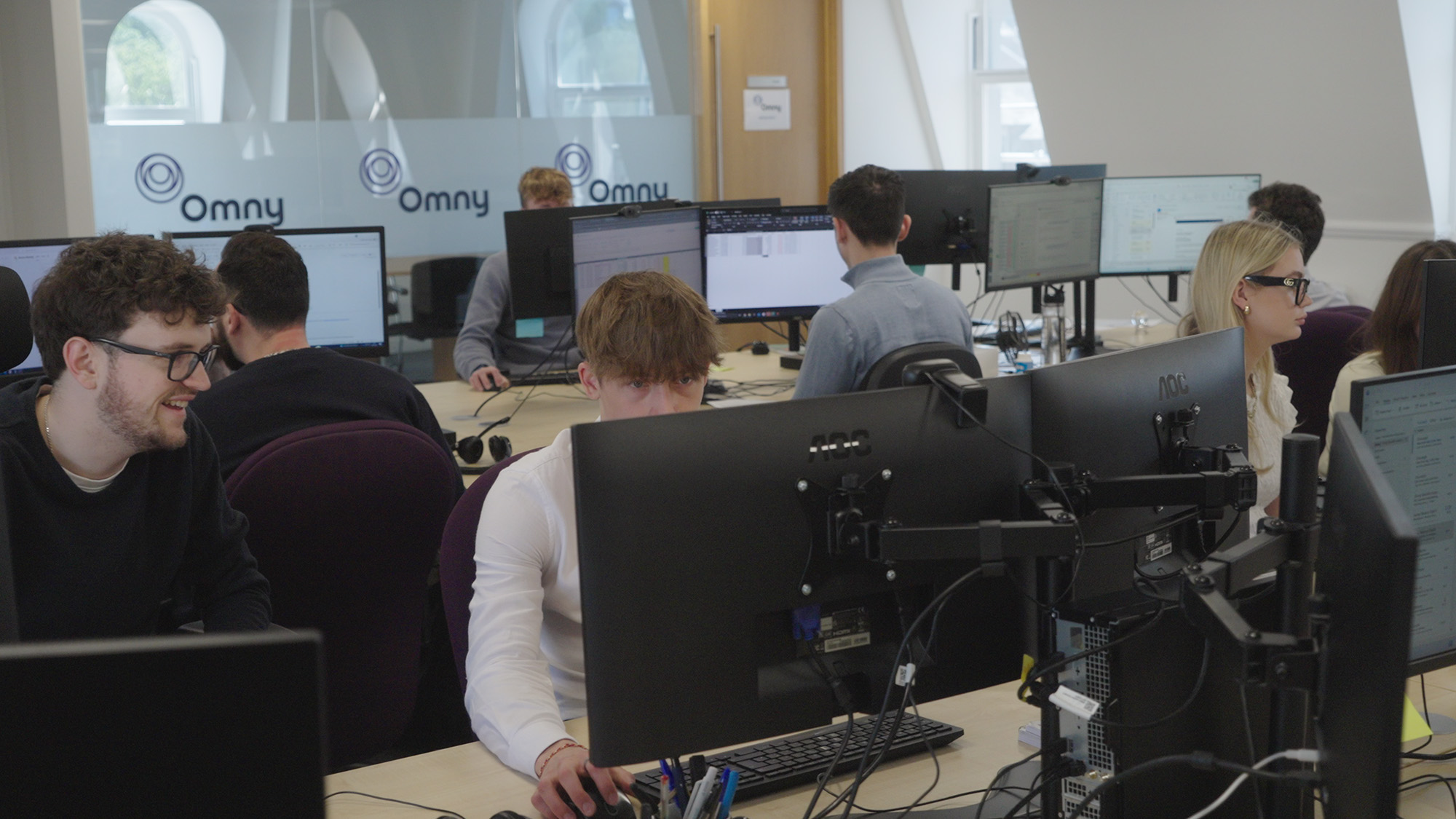 People working at computer stations in a modern office with Omny branding on the glass wall